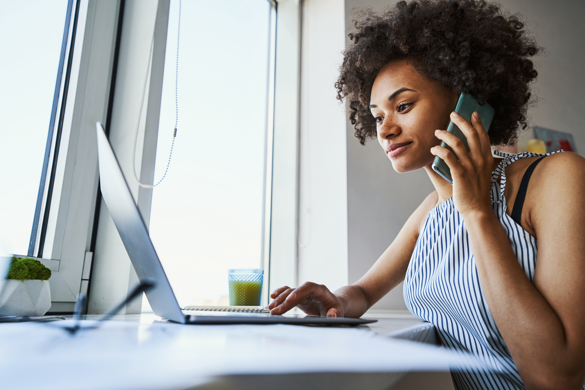 Focused beautiful young woman with a smartphone pressed to her ear sitting at her laptop