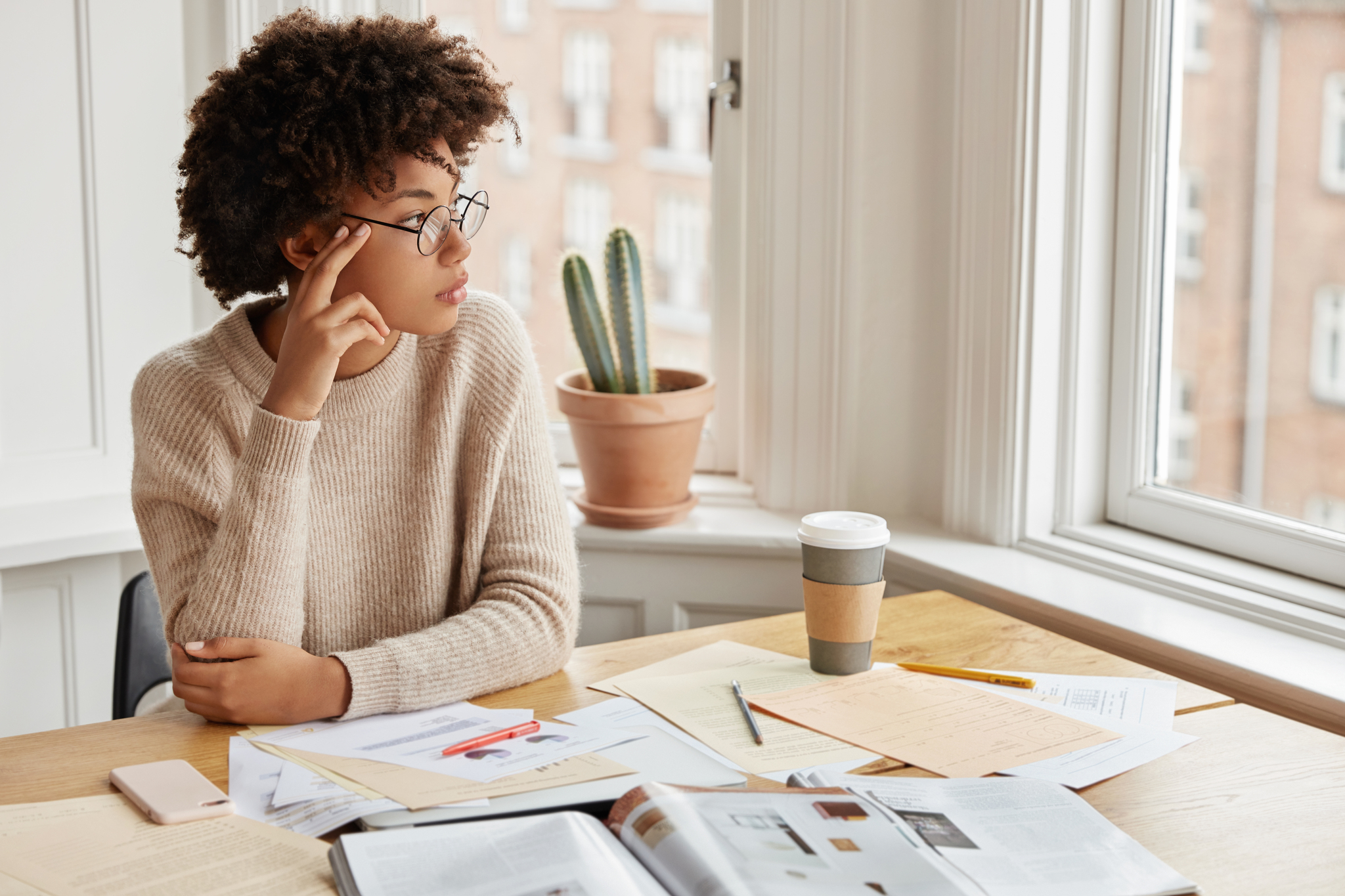 Indoor shot of pensive young African American designer surrounded with many papers and takeaway coffee, busy with work, thinks about creation new business plan, poses in cozy room near window.