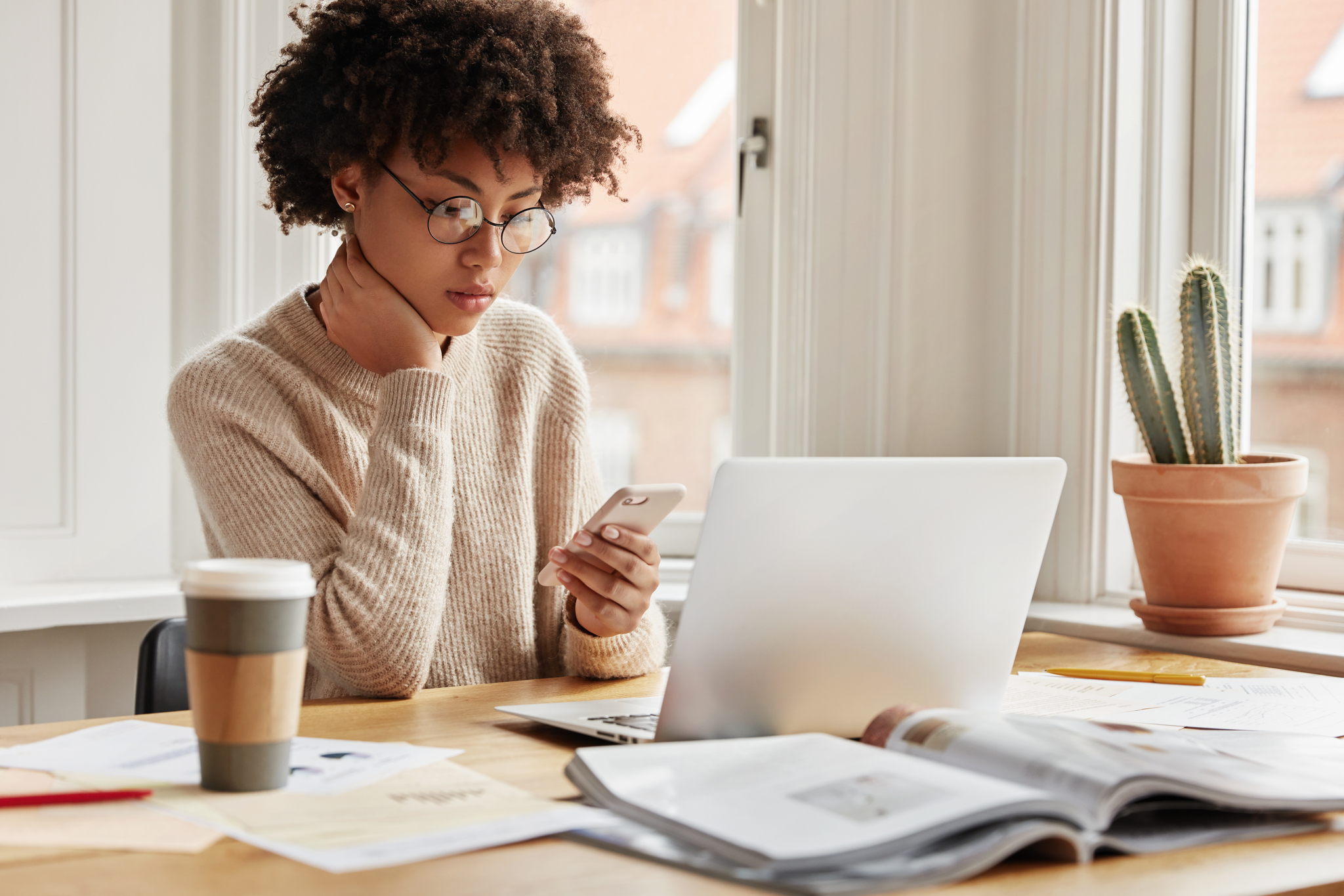 Serious female worker sits at desktop with laptop computer, surrounded with papers and journals, types sms message on smart phone device, connected to wireless internet, poses near window indoor